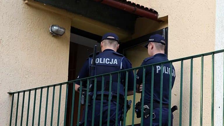Dwóch policjantów stoi przy wejściu do domu, rozmawiając, z widocznymi oznaczeniami 'Policja' na mundurach.