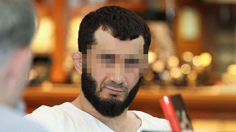Mężczyzna z ciemnym zarostem i krótkimi włosami, rozmyte tło, w trakcie rozmowy lub wywiadu.