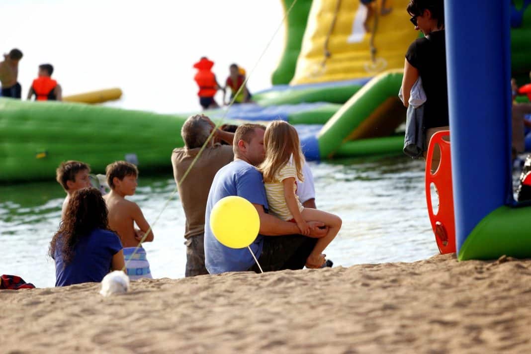 Rodzina siedzi na plaży, w tle duża zjeżdżalnia wodna na jeziorze.