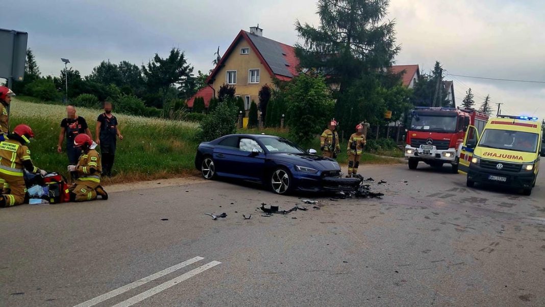 Rozbity samochód sportowy po czołowym zderzeniu na drodze, w pobliżu strażacy i służby ratunkowe.