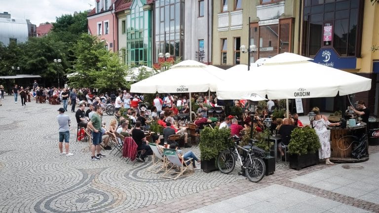 Tłum ludzi siedzących i stojących przy stolikach na rynku, pod parasolami i w cieniu drzew.