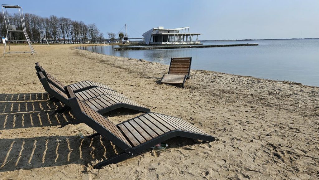 Plaża nad jeziorem z leżakami i nowoczesnym budynkiem na molo w tle.