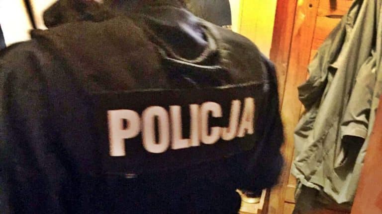 Policjant w czarnej kurtce z napisem 'POLICJA' na plecach, stojący w pomieszczeniu.