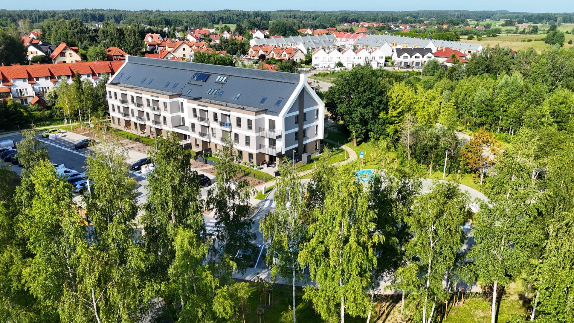 Widok na nowoczesne osiedle mieszkaniowe otoczone zielenią i drzewami, z parkingiem i placem rekreacyjnym.