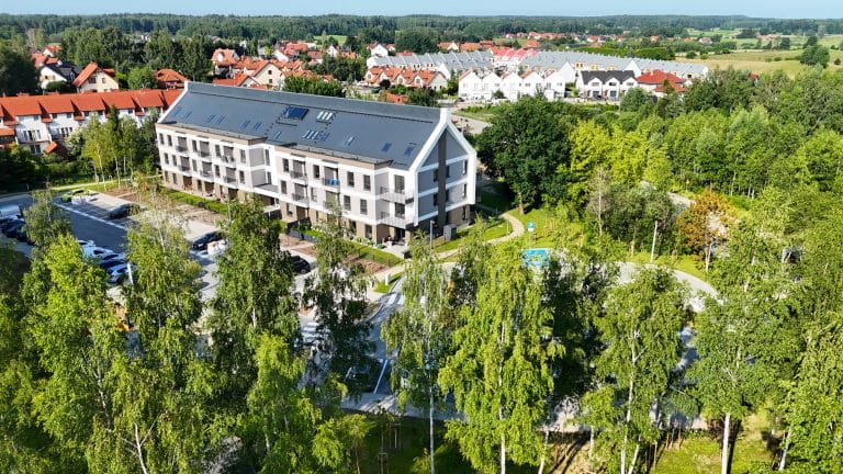 Widok na nowoczesne osiedle mieszkaniowe otoczone zielenią i drzewami, z parkingiem i placem rekreacyjnym.