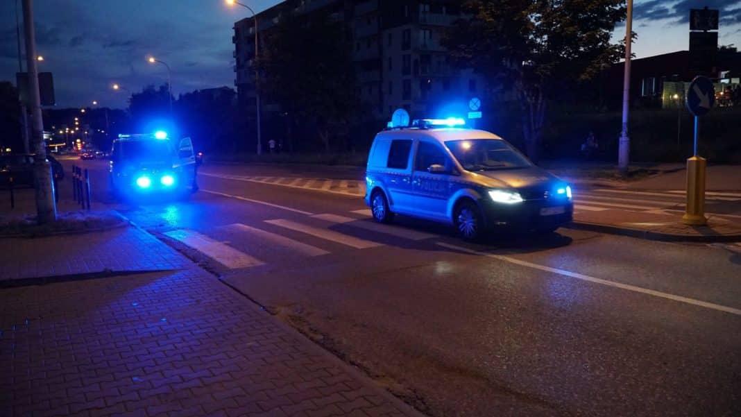 Policja zatrzymuje pojazdy na ulicy w nocy, w tle oświetlone ulice i budynki.