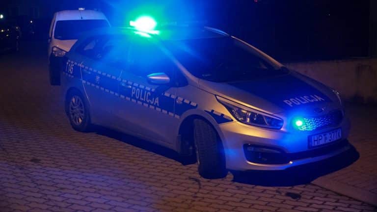 Policja na miejscu interwencji, na pierwszym planie radiowóz z włączonymi światłami, nocna scena.
