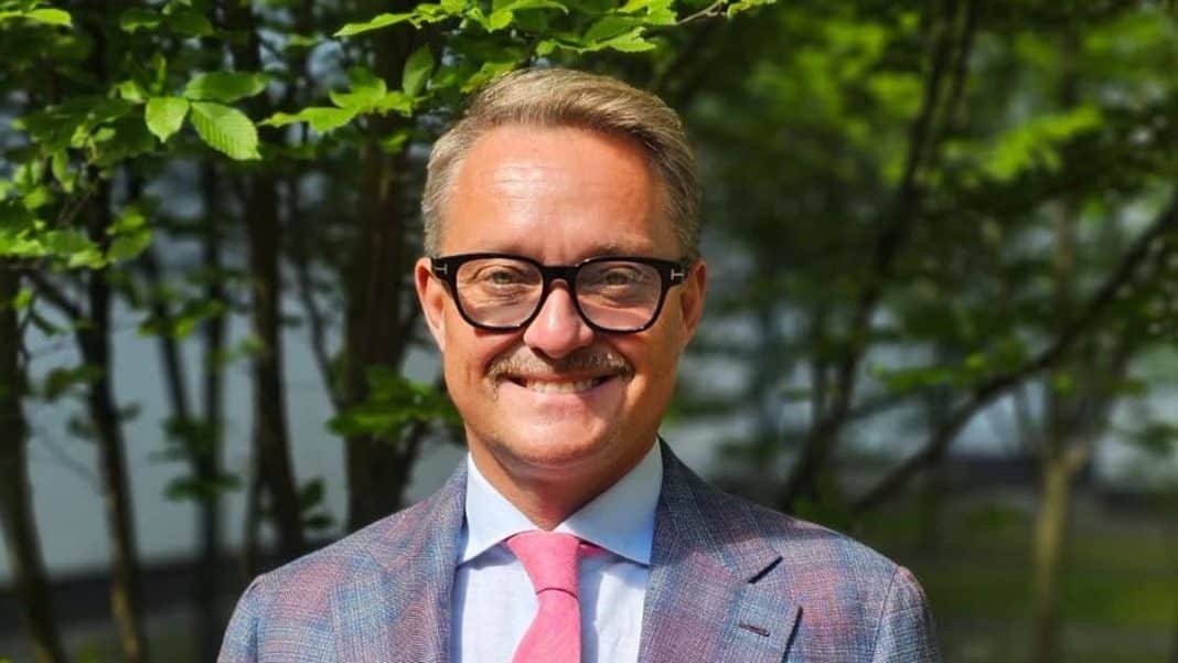 Mężczyzna w okularach i garniturze uśmiecha się na tle zielonych liści.