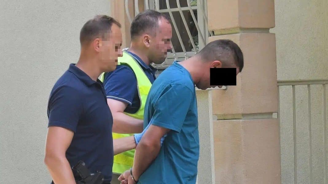 Mężczyzna w niebieskiej koszuli jest zatrzymywany przez policjantów na tle ściany budynku.