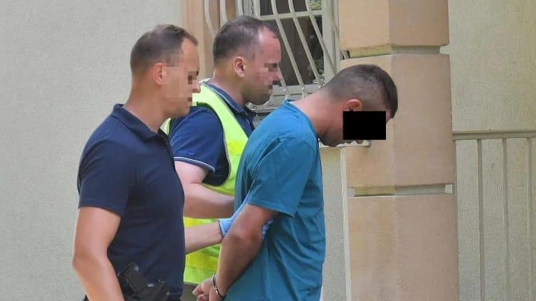 Mężczyzna w niebieskiej koszuli jest zatrzymywany przez policjantów na tle ściany budynku.