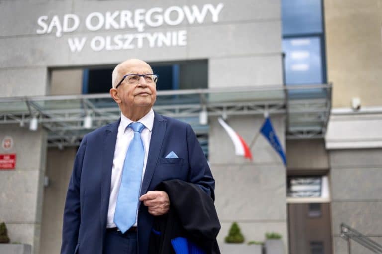 Mężczyzna w garniturze stoi przed budynkiem sądu w Olsztynie, za nim widoczny napis "Sąd Okręgowy w Olsztynie".