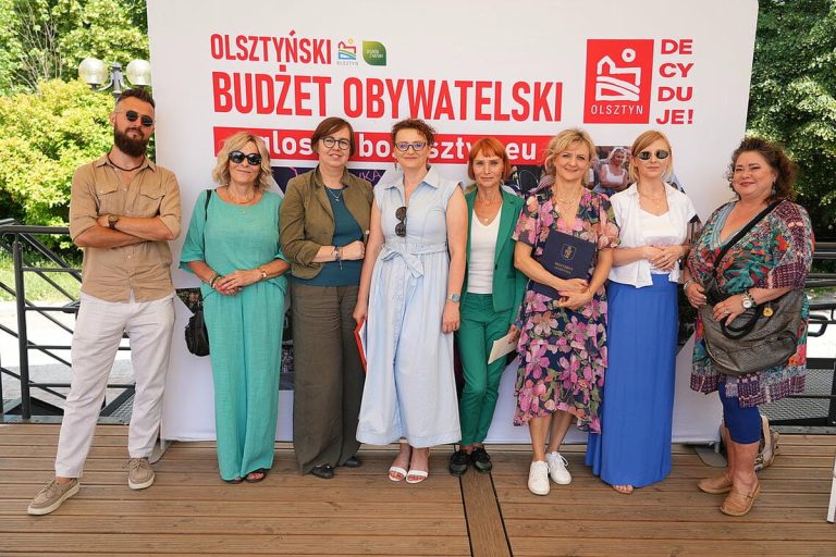 Grupa osób na tle baneru z napisem 'Olsztyński Budżet Obywatelski' podczas wydarzenia na zewnątrz.