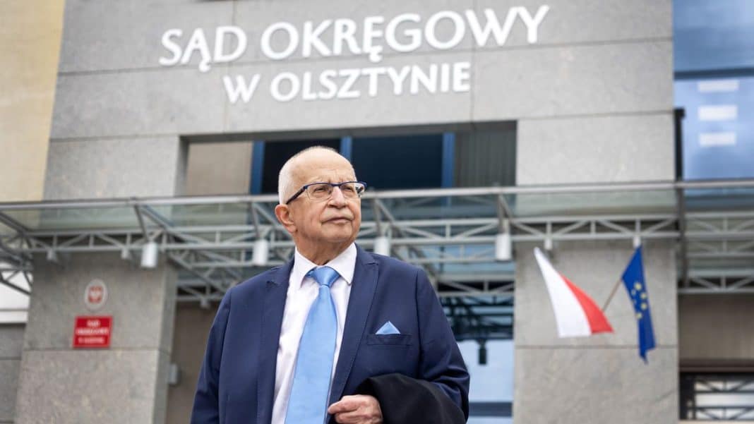 Starszy mężczyzna w garniturze i okularach stoi przed budynkiem sądu w Olsztynie.
