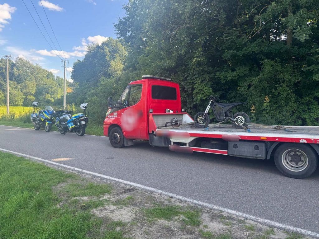 Czerwony samochód z lawetą, na której znajduje się motocykl, oraz dwa policyjne radiowozy na poboczu drogi, w tle drzewa.