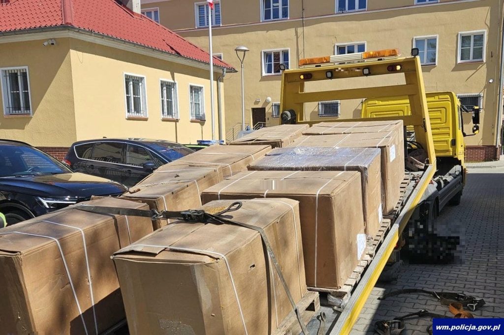 Na zdjęciu widoczny jest żółty samochód dostawczy z dużą ilością kartonów na pace, zaparkowany na ulicy w otoczeniu budynków.