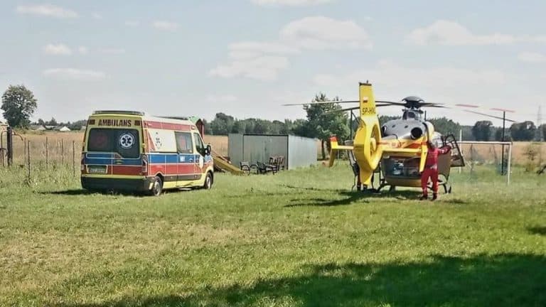 Na polu stoją śmigłowiec Lotniczego Pogotowia Ratunkowego i ambulans, w tle zielona łąka i niebo z chmurami.