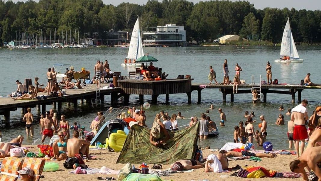 Plaża nad jeziorem z ludźmi opalającymi się i kąpiącymi w wodzie, w tle las i łodzie na wodzie.