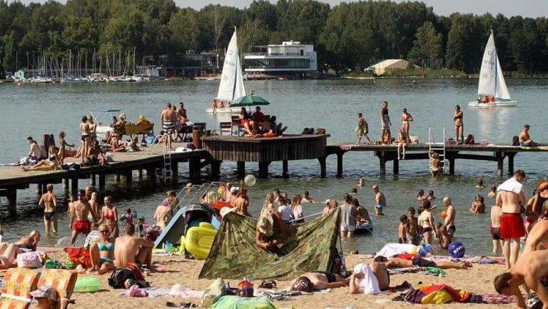 Plaża nad jeziorem z ludźmi opalającymi się i kąpiącymi w wodzie, w tle las i łodzie na wodzie.