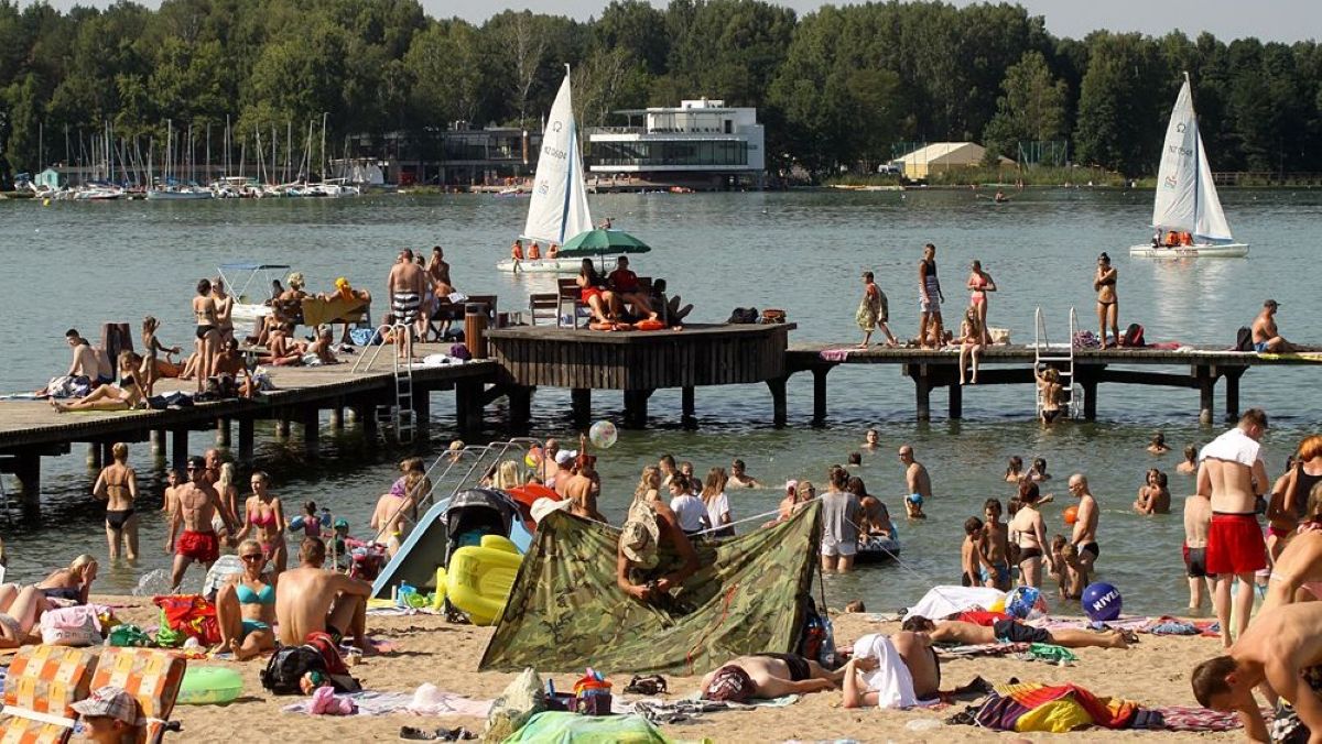 Plaża nad jeziorem z ludźmi opalającymi się i kąpiącymi w wodzie, w tle las i łodzie na wodzie.