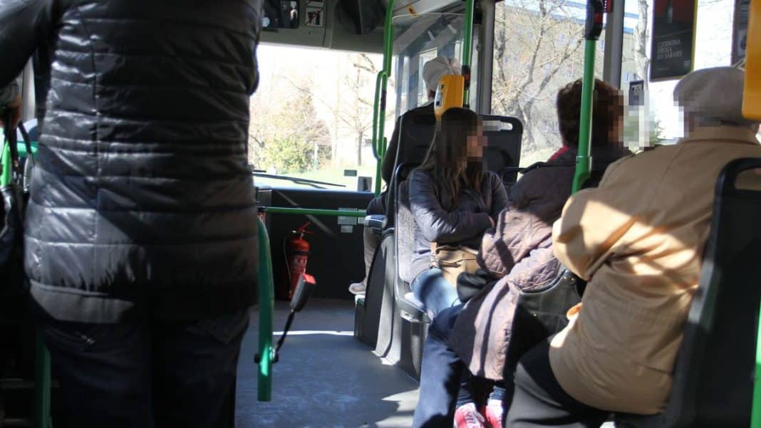 Widok wnętrza autobusu z pasażerami, w tle kierowca przy kierownicy, na zdjęciu widoczne osoby siedzące i stojące.