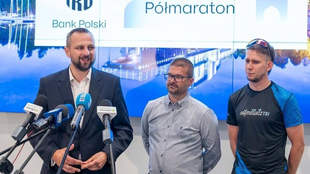 Trzech mężczyzn podczas konferencji prasowej z mikrofonami i ekranem w tle.