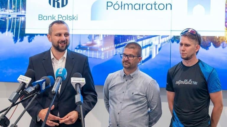 Trzech mężczyzn podczas konferencji prasowej z mikrofonami i ekranem w tle.
