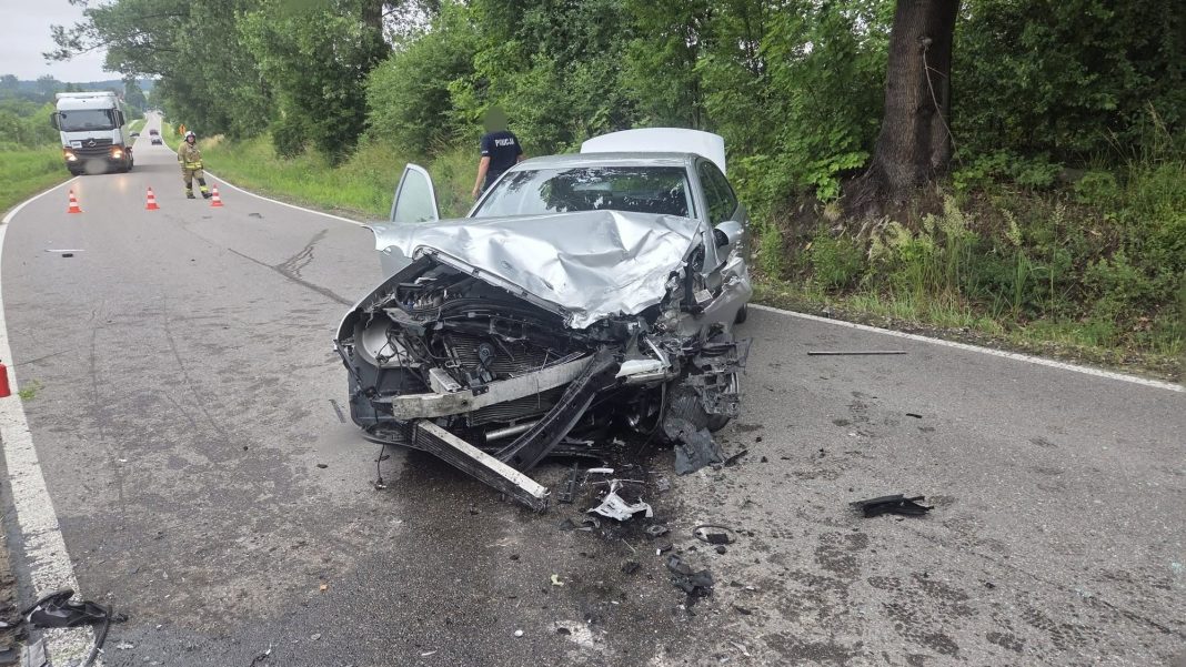 Rozbity samochód po zderzeniu na drodze w Tomaszkowie, w tle strażacy i policja.