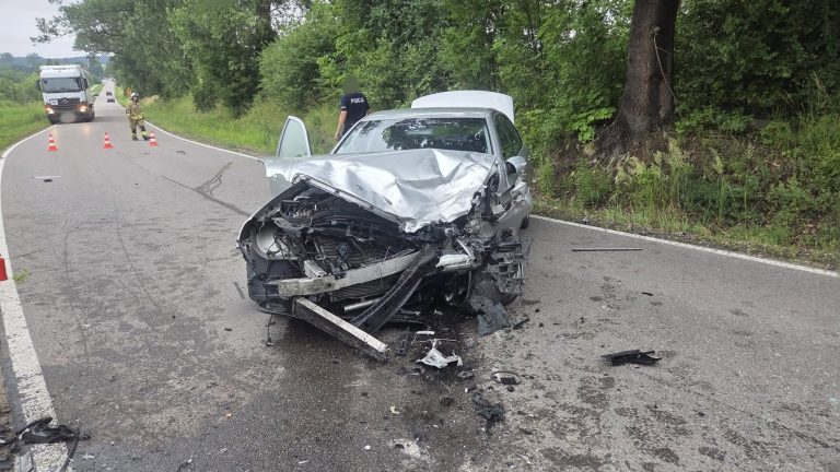 Rozbity samochód po zderzeniu na drodze w Tomaszkowie, w tle strażacy i policja.