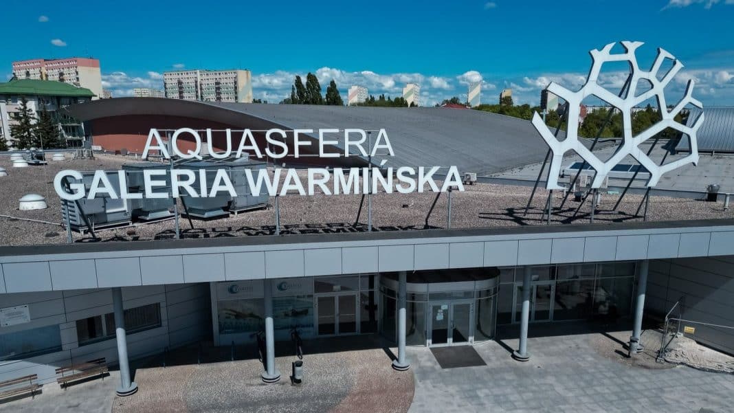 Widok na zewnętrzną część basenu Aquasfera w Olsztynie, z dużym napisem na dachu i nowoczesną architekturą.