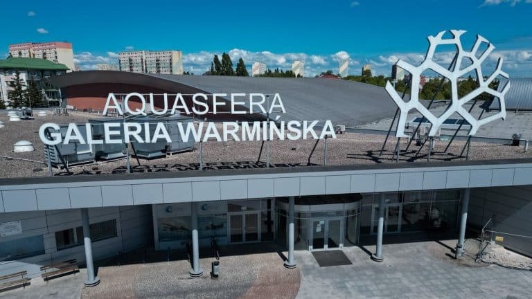 Widok na zewnętrzną część basenu Aquasfera w Olsztynie, z dużym napisem na dachu i nowoczesną architekturą.