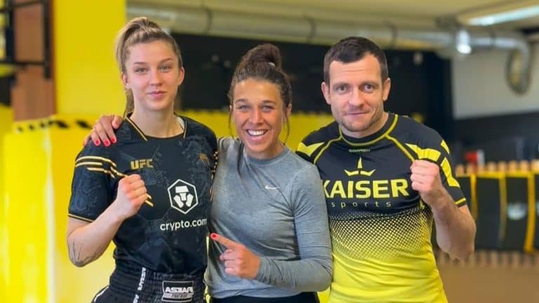 Trzy osoby w sportowych strojach, jedna z nich to zawodniczka MMA, na tle sali treningowej.