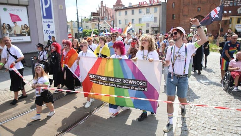 Uczestnicy Marszu Równości maszerują z transparentem w centrum Olsztyna, w tłumie ludzi i flag.