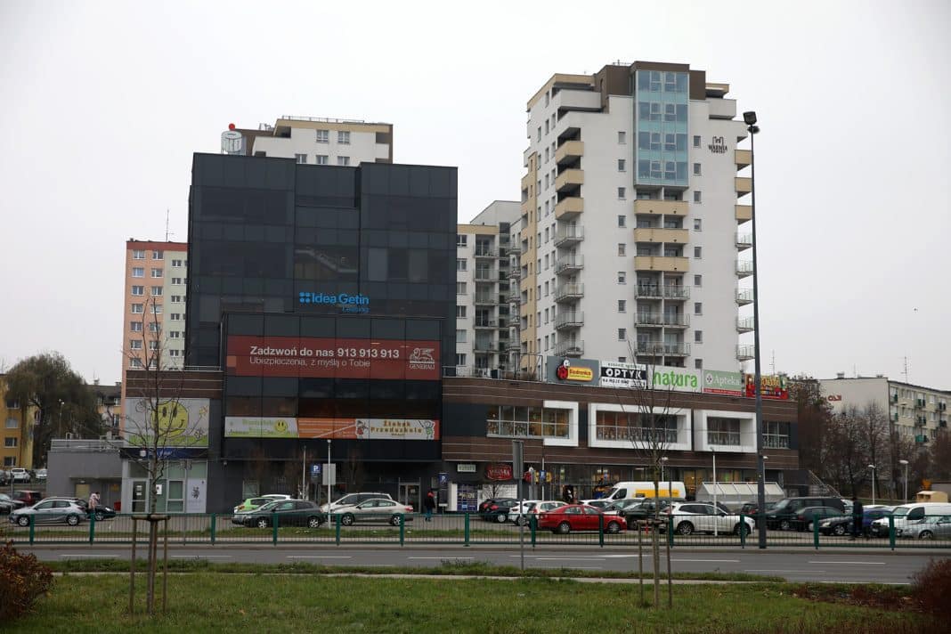Widok na wielopiętrowy budynek z sklepami i parkingiem przed nim, w tle szare niebo.