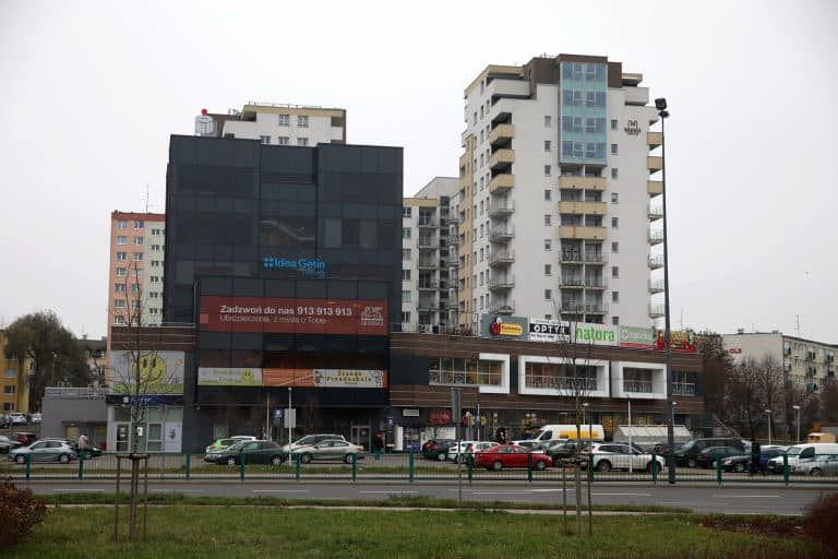 Widok na wielopiętrowy budynek z sklepami i parkingiem przed nim, w tle szare niebo.