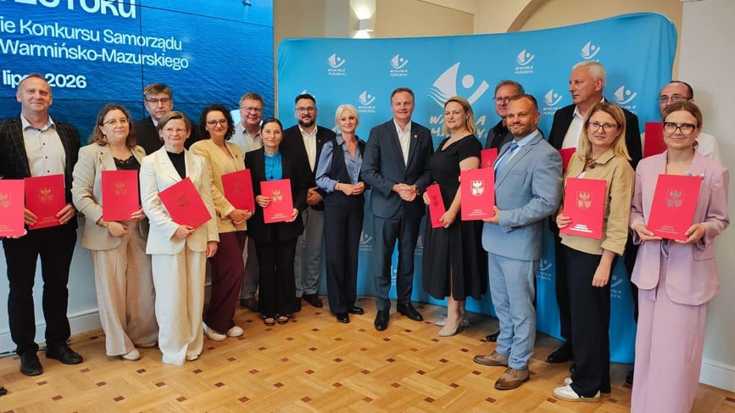 Grupa osób na oficjalnej uroczystości z czerwonymi teczkami, w tle logo i napis o konkursie na Warmii i Mazurach.