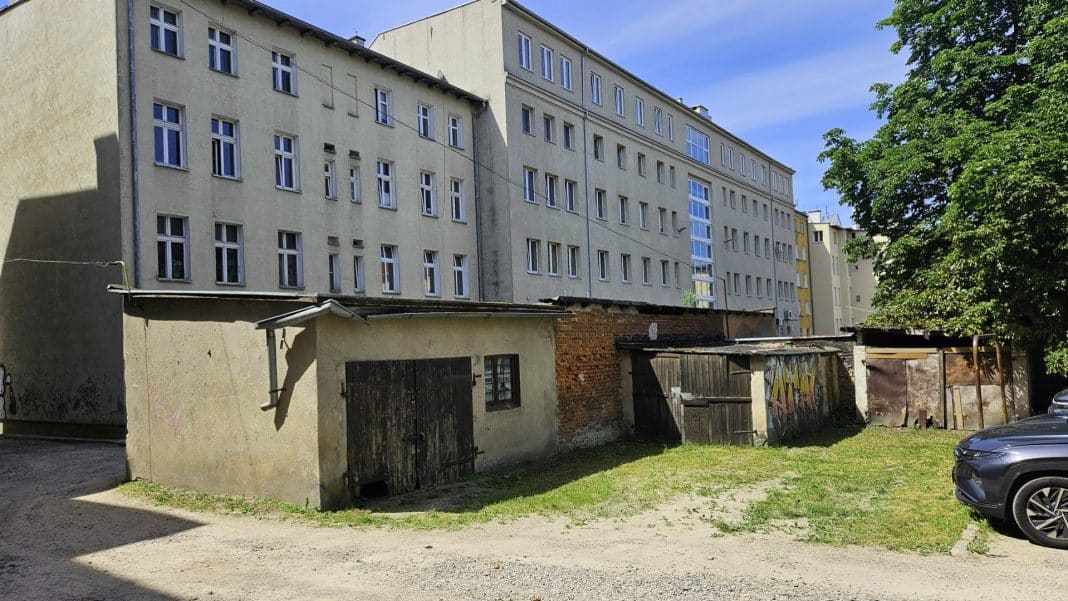Stare budynki gospodarcze i garaże w centrum Olsztyna, na tle nowoczesnego bloku mieszkalnego.