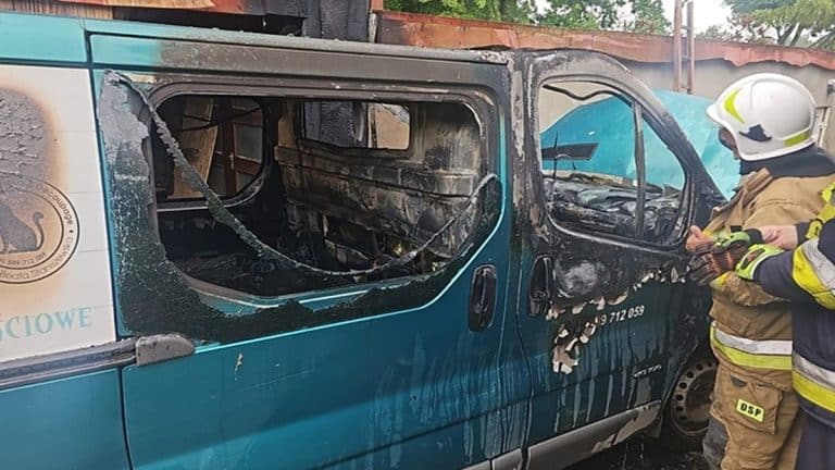 Spalony samochód dostawczy i sprzęt ratowniczy po pożarze, strażak w hełmie i odzieży ochronnej przy pojeździe.