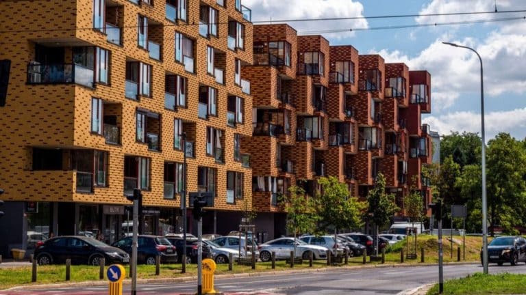 Nowoczesny blok mieszkalny z wieloma balkonami i parkingiem przed budynkiem.