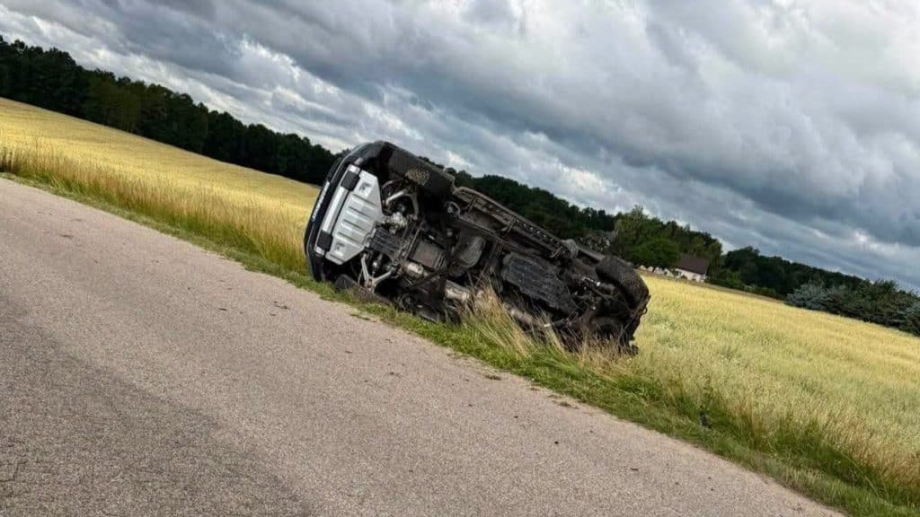 Rozbity samochód leży na boku na poboczu w polu, nad nim ciemne chmury na niebie.