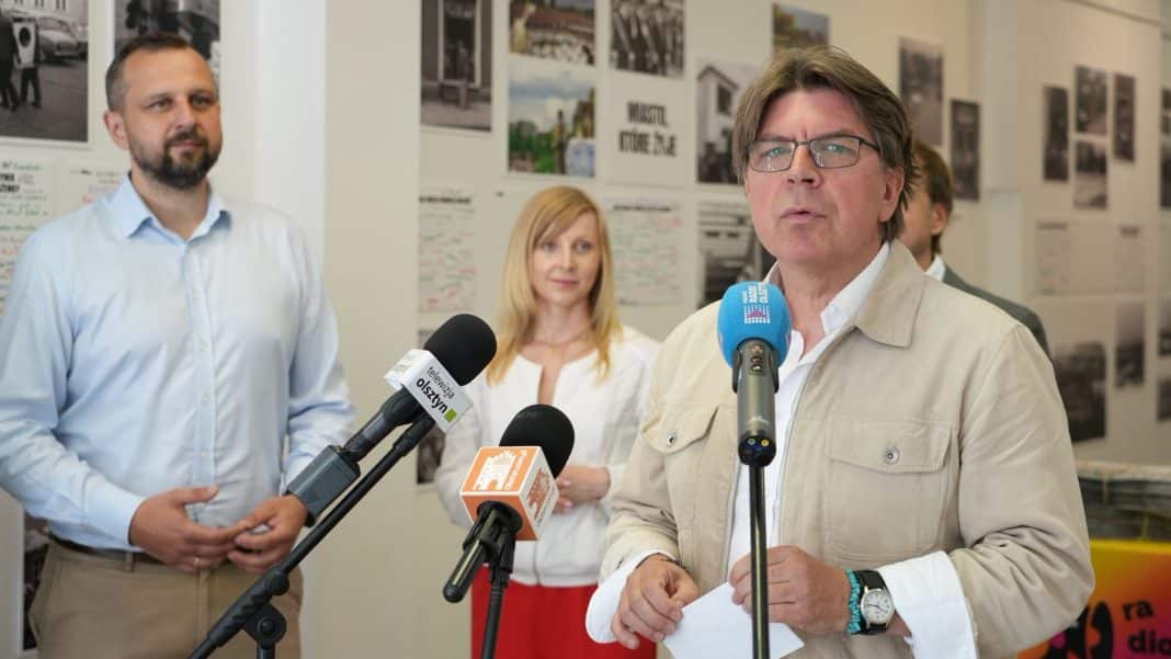 Mężczyzna w okularach i jasnej kurtce mówi do mikrofonów podczas konferencji, w tle kobieta i mężczyzna przy ścianie z fotografiami.