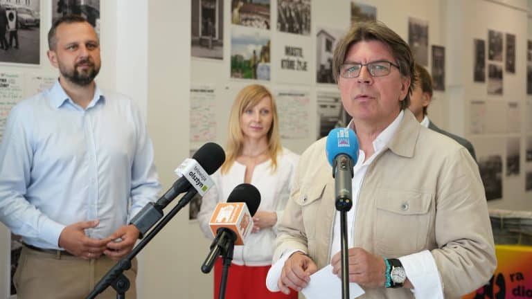 Mężczyzna w okularach i jasnej kurtce mówi do mikrofonów podczas konferencji, w tle kobieta i mężczyzna przy ścianie z fotografiami.