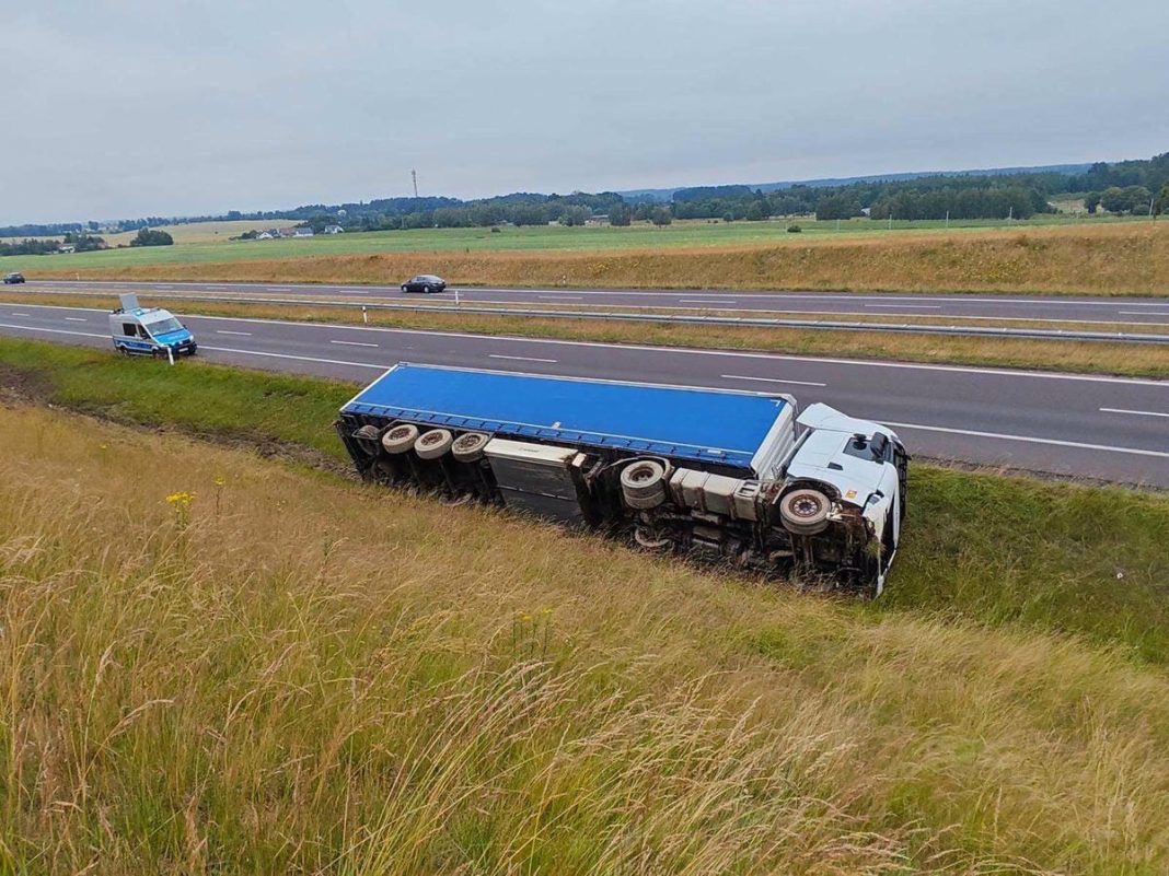 Ciężarówka przewrócona na boku w rowie przy drodze S7, w pobliżu pasa ruchu, z widocznymi pojazdami i terenami zielonymi w tle.