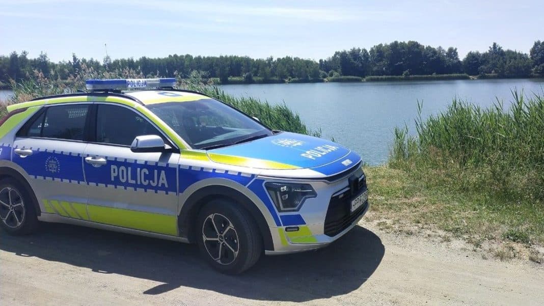Policja na brzegu jeziora, w tle roślinność i woda, w słoneczny dzień.
