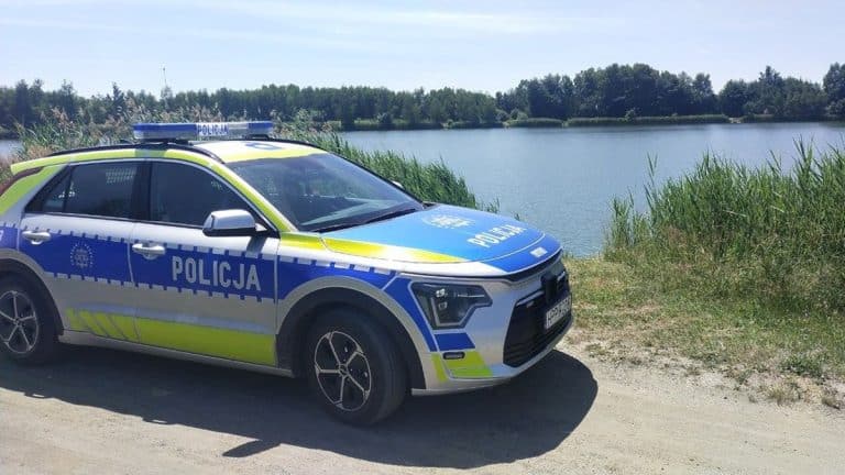 Policja na brzegu jeziora, w tle roślinność i woda, w słoneczny dzień.