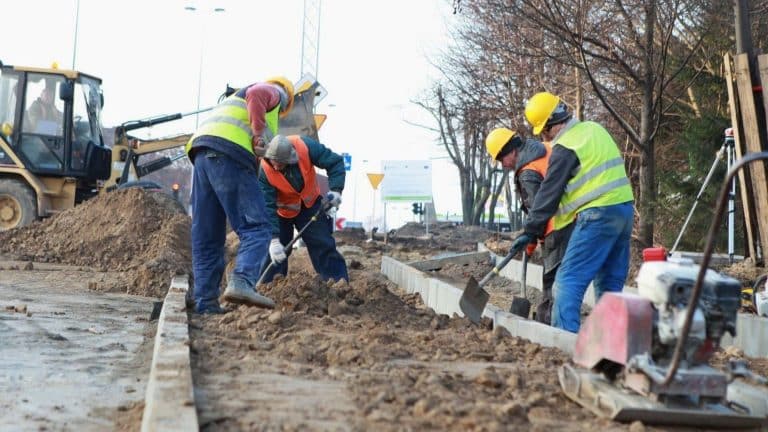 Pracownicy budowlani w odblaskowych kamizelkach i kaskach pracują na placu budowy, kopiąc ziemię.