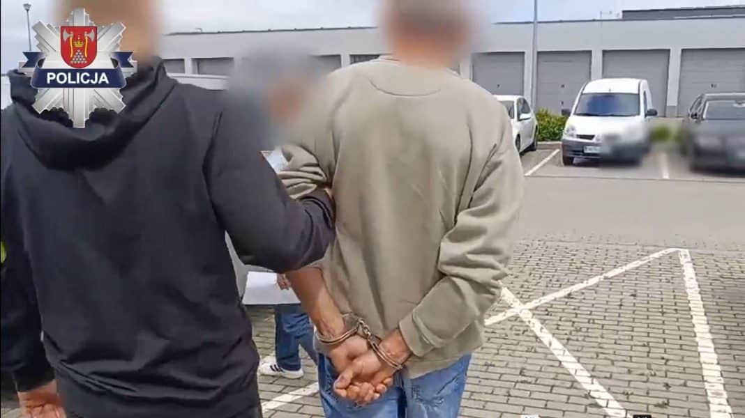 Mężczyzna w rękawicach z kajdankami na nadgarstkach, otoczony przez policjantów na parkingu.