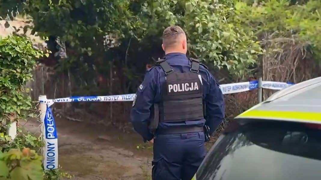 Policjant stoi na miejscu akcji, otoczony taśmą policyjną, w tle zarośla i samochód służbowy.