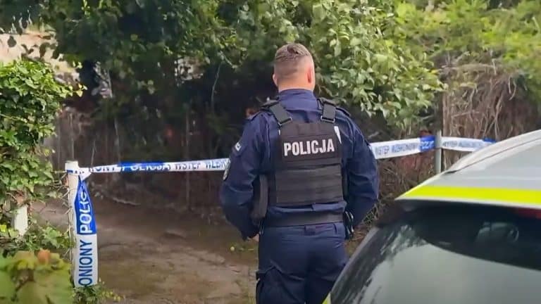 Policjant stoi na miejscu akcji, otoczony taśmą policyjną, w tle zarośla i samochód służbowy.