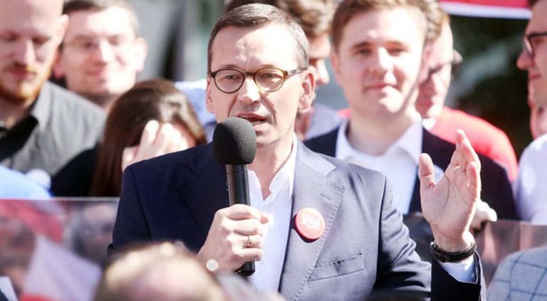 Mężczyzna w okularach i garniturze przemawia do zgromadzonej grupy ludzi, trzymając mikrofon.