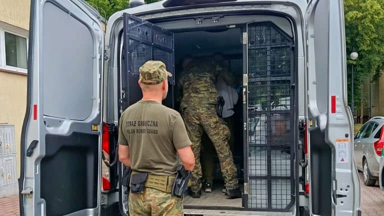 Policjant i funkcjonariusze Straży Granicznej w pojeździe, w tle drzewa i budynki.